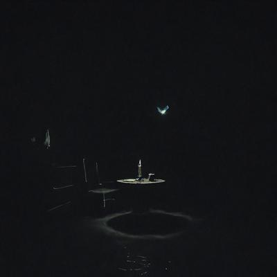 The primary subject of the image is a lit candle on a table. The setting is a dark room, and the visual style appears to be a photo. The dominant colors are black and white, creating a moody atmosphere.